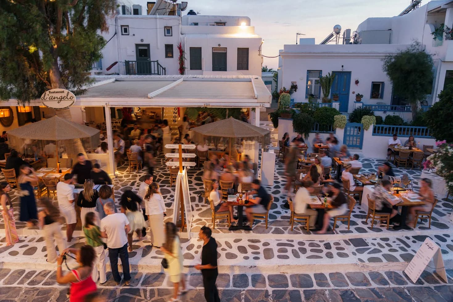 d'angelo restaurant terrace at dusk in Mykonos Town with guests dining under the iconic tree and restaurant sign
